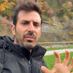 Oratore all'aperto che spiega con una mano in evidenza, sfondo di alberi autunnali e prato.