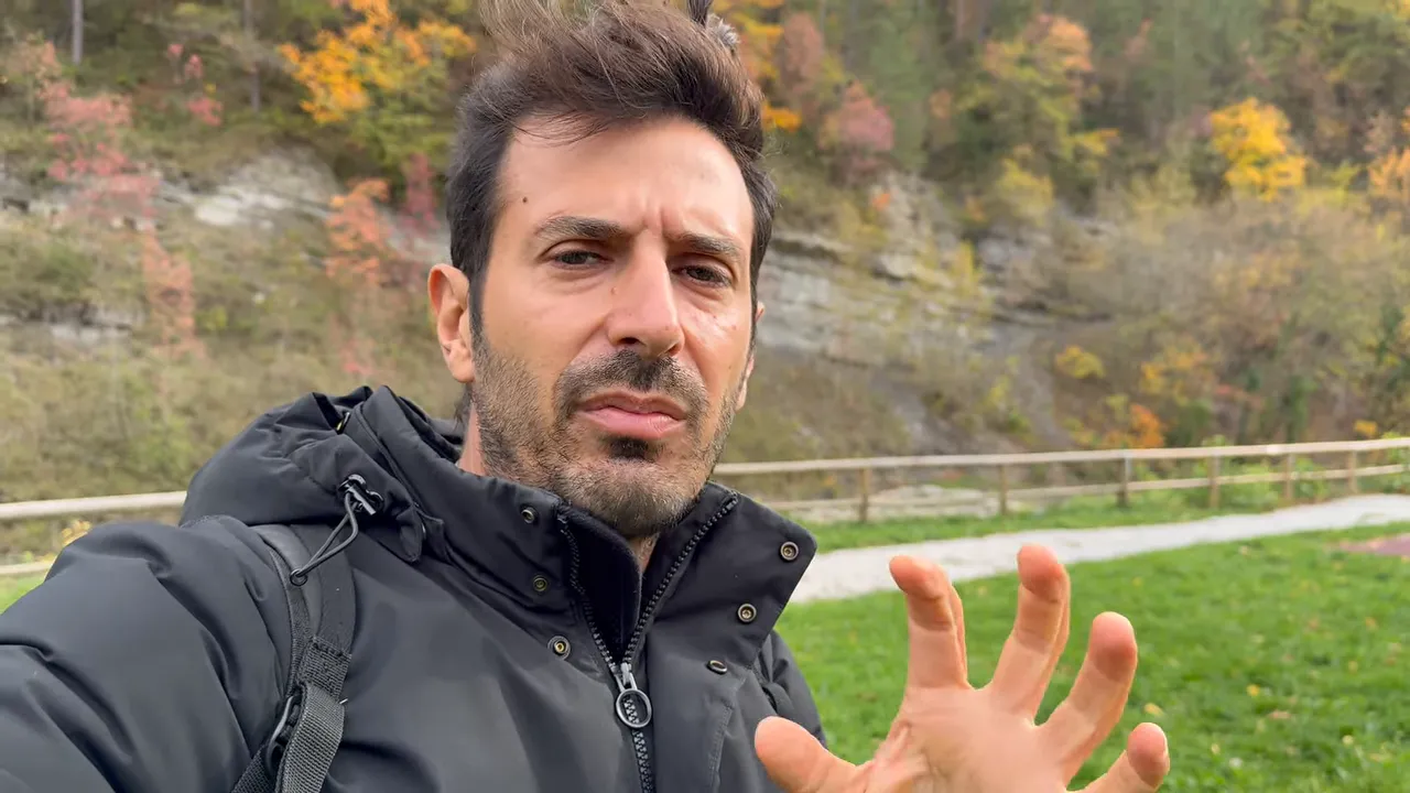 Oratore all'aperto che spiega con una mano in evidenza, sfondo di alberi autunnali e prato.