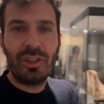 Primo piano di una persona con una teca espositiva e un sarcofago antico visibili sullo sfondo