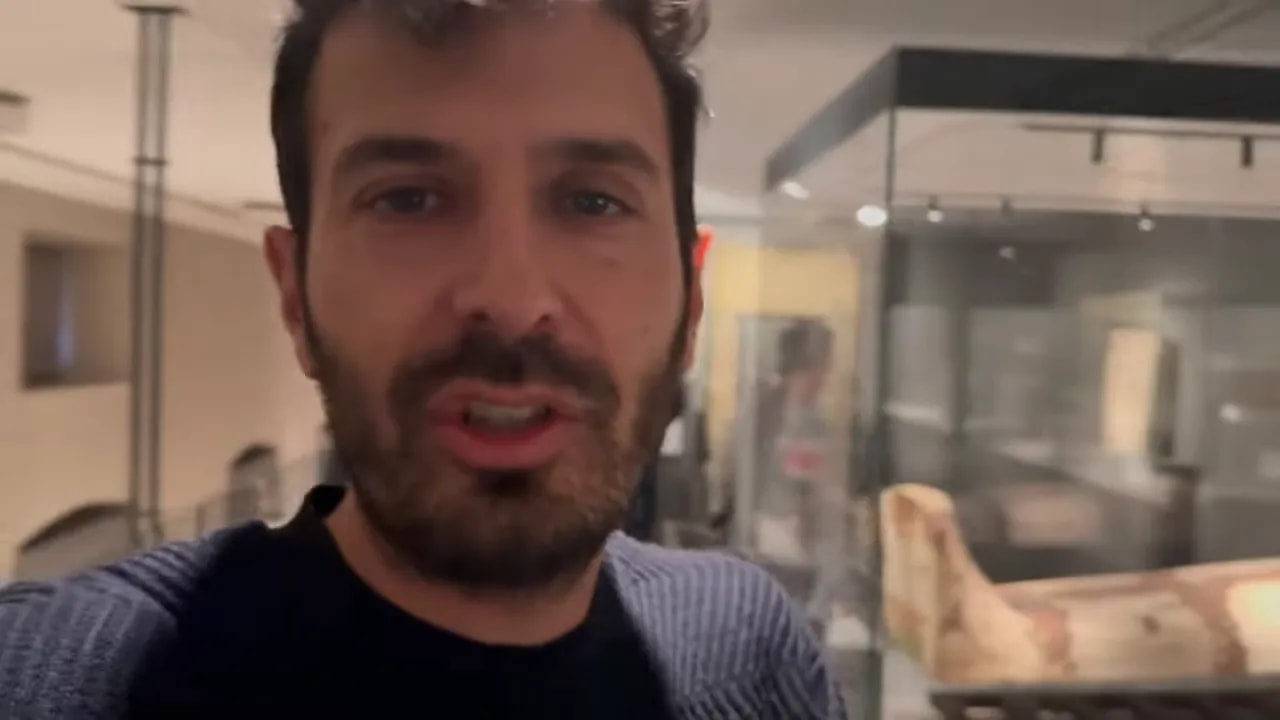 Primo piano di una persona con una teca espositiva e un sarcofago antico visibili sullo sfondo